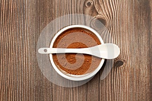 Cocoa powder in a white bowl with a white spoon on it and on a wooden background