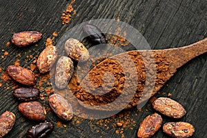 Cocoa powder on a wooden spoon isolated on black rustic wood board from above. Spilt cocoa and roasted peeled and unpeeled cocoa