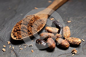 Cocoa powder on a wooden spoon next to roasted peeled and unpeeled cocoa beans and chocolate shavings on black slate