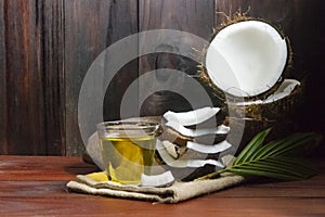 Coconut oil with coconut half and coconut pieces and leaf on wooden table and black wooden background.