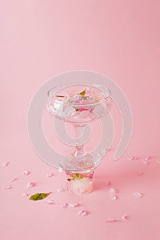 Coctail glass with rose petals standing on ice cube with a flower on pink background