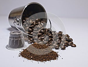 Coffee beans fallen out of silver bucket with ground coffee and pod on white background