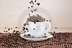 Coffee beans falling down into white cup standing on white plate standing on tablemat.