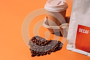 Coffee beans in the shape of a heart on an orange background next to packaging with text decaf and a paper cup in a tray
