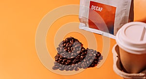 Coffee beans in the shape of a heart on an orange background next to packaging with text decaf and a paper cup in a tray