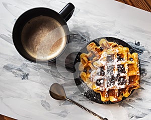Coffee in a black tall black mug and waffles sprinkled with icing sugar