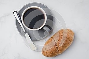 Coffee break with croissant butter knife and cup of black coffee on a white stone table. Top view