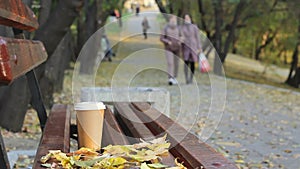 Coffee break on empty bench in autumn city park, blurred two woman at public park walkway on background