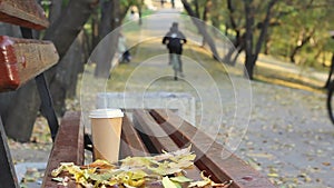 Coffee break on empty bench in autumn city park, two blurred boys on a bicycles at public park walkway on background