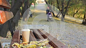 Coffee break on empty bench in autumn city park, woman with baby carriage at public park walkway on background