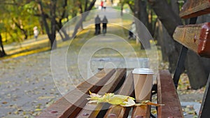 Coffee break on empty bench in autumn park, blurred couple at walkway on backgr