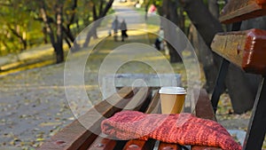 Coffee break on empty bench in autumn park, blurred couple at walkway on backgr