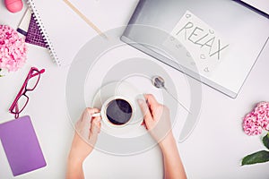 Coffee break time on the workspace - female hands with cup of coffee on the white working office desk with laptop, notebook, glass