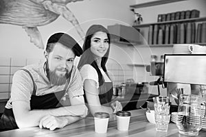 Coffee Business Concept - Positive young bearded man and beautiful attractive lady barista couple in apron looking at