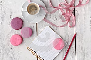 Coffee, cake macaron, clean notebook, eyeglasses and flower on pink table from above. Female working desk. Cozy breakfast. Flat la