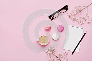 Coffee, cake macaron, notebook, eyeglasses and flower on pink table from above. Female working desk. Cozy breakfast. Flat lay.