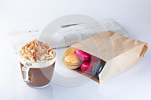 Coffee, cappuccino in a glass mug with whipped cream and ground nut. Macaron cakes in a paper bag, newspaper.