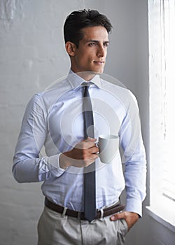 Coffee and contemplation. A handsome young businessman having a cup of coffee and looking out a window.