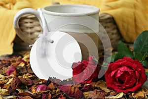 Coffee cup with blank tag label paper, two red roses and rattan basket on dried rose petals background. Still life concept..