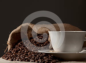 Coffee cup with burlap sack of roasted beans against dark wood b