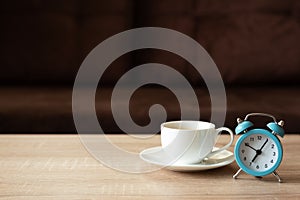 Coffee cup and clock on wooden table against defocused sofa with pillows. Front view. Good morning concept. Mock-up