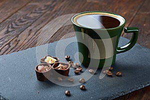 Coffee cup, coffee beans, chocolate candies on stone board over wooden background, selective focus, close-up