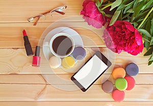 Coffee cup with colorful macaroons, smart phone, lipstck, nail polish and red peony flowers on wooden table.