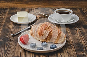 Coffee cup, croissant with berries in white bowl and butter knife on wooden table. Selective focus. Healthy breakfast with fresh