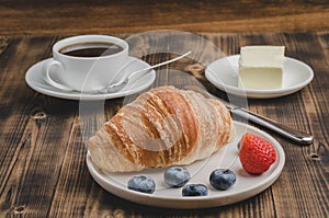 Coffee cup, croissant with berries in white bowl and butter knife on wooden table. Selective focus. Healthy breakfast with fresh