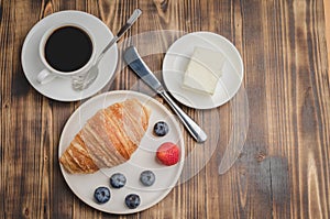 Coffee cup, croissant with berries in white bowl and butter knife on wooden table. Top view and copyspace. Healthy breakfast with