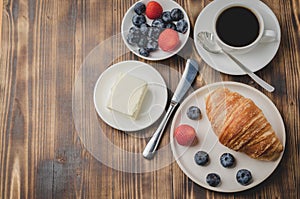 Coffee cup, croissant with berries in white bowl and butter knife on wooden table. Top view and copyspace. Healthy breakfast with