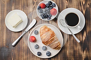 Coffee cup, croissant with berries in white bowl and butter knife on wooden table. Top view. Healthy breakfast with fresh berries