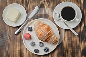 Coffee cup, croissant with berries in white bowl and butter knife on wooden table. Top view. Healthy breakfast with fresh berries