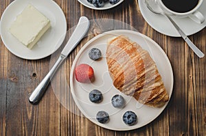 Coffee cup, croissant with berries in white bowl and butter knife on wooden table. Top view. Healthy breakfast with fresh berries