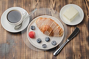 Coffee cup, croissant with berries in white bowl and butter knife on wooden table. Top view. Healthy breakfast with fresh berries