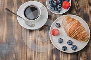Coffee cup, croissant with berries in white bowl and butter knife on wooden table. Top view. Healthy breakfast with fresh berries