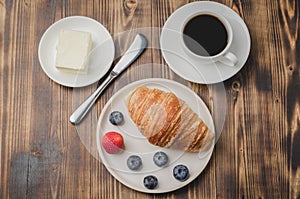 Coffee cup, croissant with berries in white bowl and butter knife on wooden table. Top view. Healthy breakfast with fresh berries