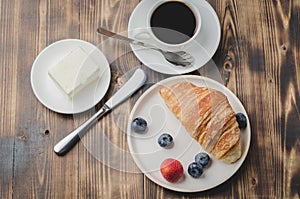 Coffee cup, croissant with berries in white bowl and butter knife on wooden table. Top view. Healthy breakfast with fresh berries