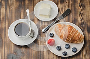 Coffee cup, croissant with berries in white bowl and butter knife on wooden table. Top view. Healthy breakfast with fresh berries