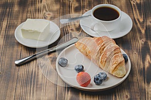 Coffee cup, croissant with berries in white bowl and butter on wooden background. Healthy breakfast with fresh berries