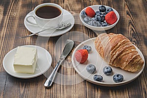 Coffee cup, croissant with berries in white bowl and butter on wooden background. Healthy breakfast with fresh berries