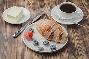 Coffee cup, croissant with berries in white bowl and butter on wooden background. Healthy breakfast with fresh berries