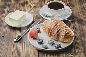 Coffee cup, croissant with berries in white bowl and butter on wooden background. Healthy breakfast with fresh berries