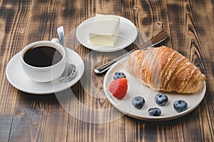 Coffee cup, croissant with berries in white bowl and butter on wooden background. Healthy breakfast with fresh berries