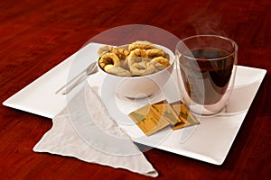 Coffee cup, double glass coffee cup with bowl of cookies, spoon, napkin and sugar sachets on white ceramic tray on wooden table un