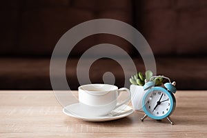 Coffee cup, flower pot and clock on wooden table against defocused sofa with pillows. Front view. Good morning concept. Mock-up