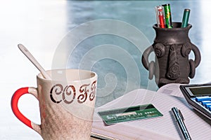 A coffee cup with a glass spoon and diary, debit card, digital calculator, a pen, and a soil pottery pen stand on the office table