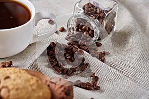 Coffee cup, jar with coffee beans, cookies over homespun tablecloth, selective focus, close-up, side view