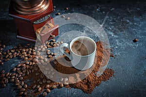 Coffee cup with steaming hot drink on ground coffee and roasted beans in front of a vintage grinder, dark background with copy