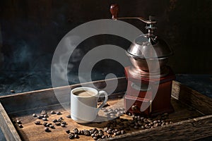 Coffee cup with steaming hot drink, nostalgic grinder and some roasted beans on a rustic wooden tray, dark background with copy